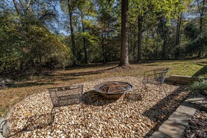 A fire pit surrounded by pebbles, a metal chair, and a metal table in a wooded area. - Modern 3 Bedroom River Retreat in Heart of Athens (Athens)