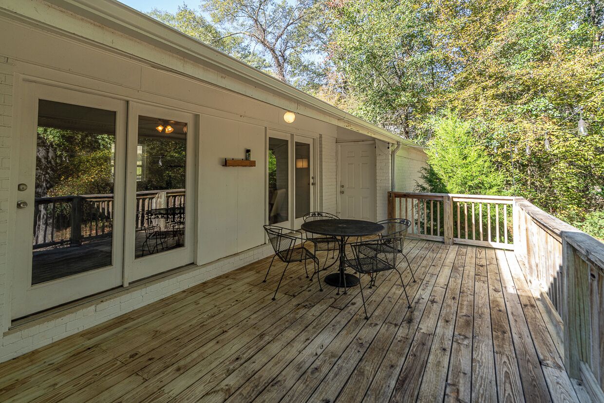 A covered outdoor deck with a round table and chairs, wooden railings, and a view of trees.