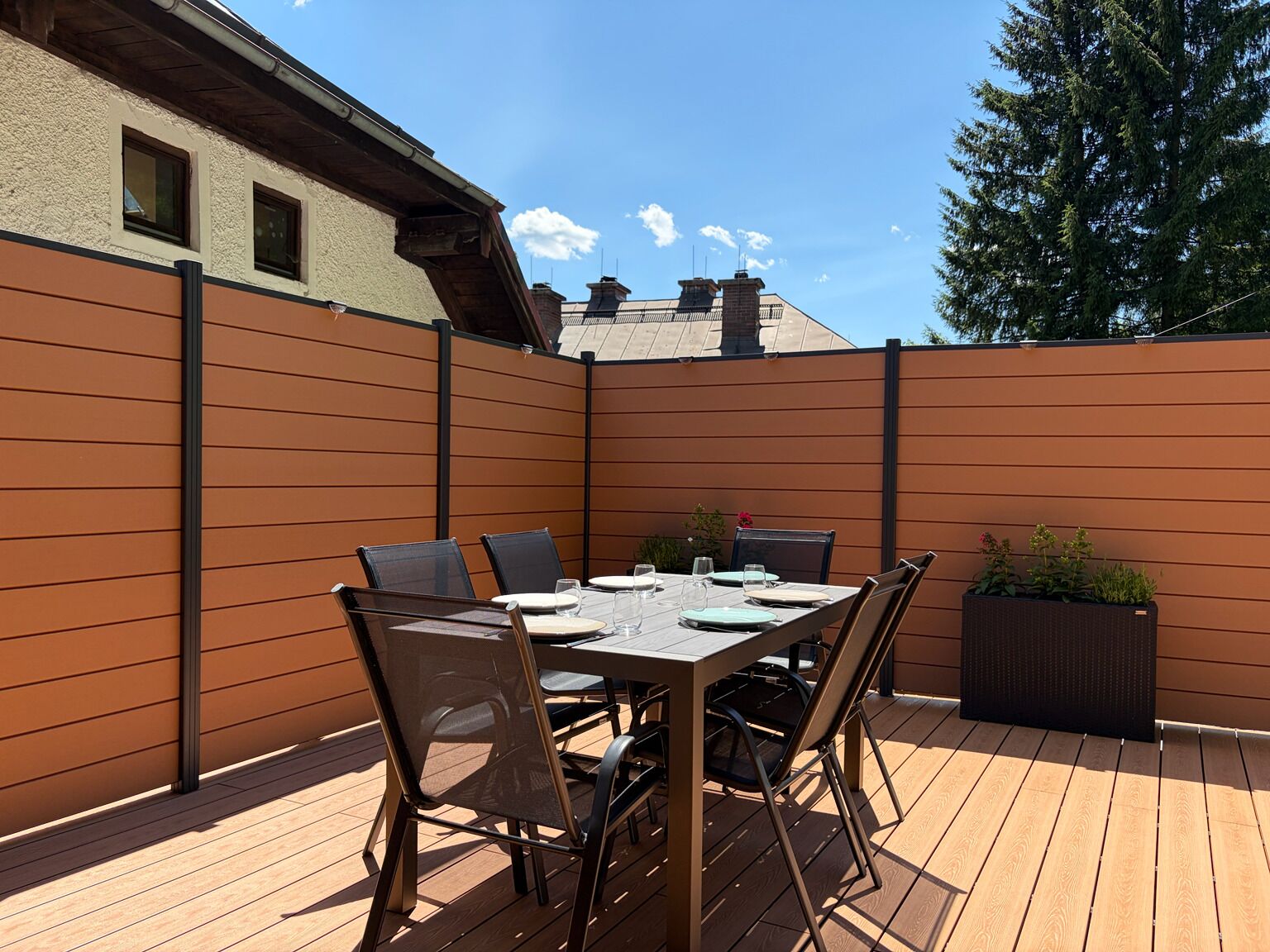 Une terrasse en bois avec une table et des chaises, une clôture et un bâtiment en arrière-plan.