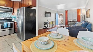 Eine moderne Küche mit Edelstahlgeräten, ein Essbereich mit Holztisch und -stühlen sowie ein Wohnzimmer mit Sofa und Fernseher.