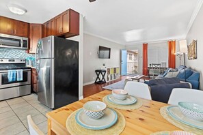 Eine moderne Küche mit Edelstahlgeräten, ein Essbereich mit Holztisch und -stühlen sowie ein Wohnzimmer mit Sofa und Fernseher.