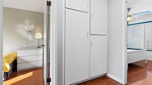 Ein Zimmer mit Holzboden, einem weißen Kleiderschrank, einem Nachttisch mit Lampe und einem Fenster mit Jalousien.