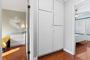 Ein Zimmer mit Holzboden, einem weißen Kleiderschrank, einem Nachttisch mit Lampe und einem Fenster mit Jalousien.