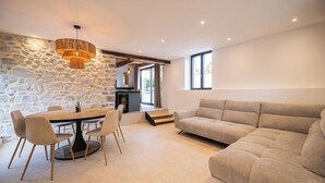 A modern living space with a stone wall, a dining area with a round table and chairs, a sofa, and a kitchen visible through an open door. - The Ostalet in Sampzon  (Sampzon)