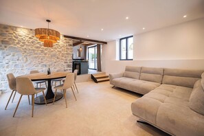 Ein modernes Wohnzimmer mit Steinwand, einem Essbereich mit rundem Tisch und Stühlen, einer Couch und einer durchgehenden Tür erkennbaren Küche.