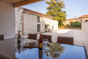 An outdoor dining area with a stone building in the background, a table with chairs, and a bottle of wine.