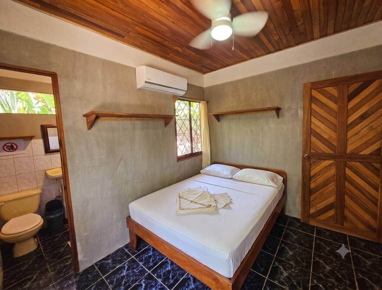 Una habitación con una cama, ventilador de techo, ventana y puerta de madera.