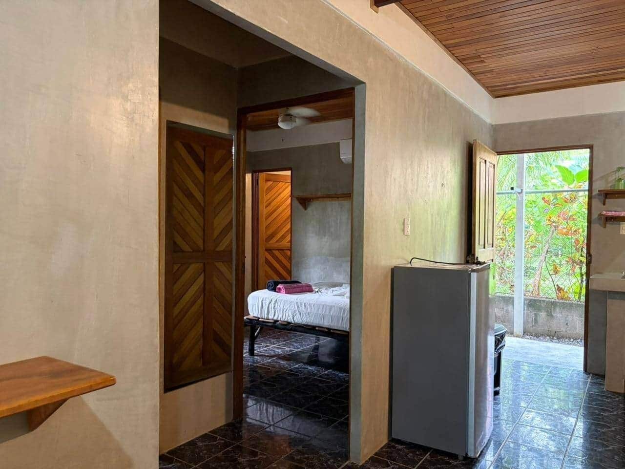 A room with a bed, a small table, a refrigerator, and a view of greenery outside.