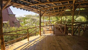 Una terraza de madera con barandal metálico, rodeada de abundante vegetación y un edificio con tejado rojo.
