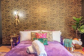 Ein Zimmer mit Wandmuster, einem Bett mit vielen Kissen, einem kleinen runden Tisch und einer dekorativen Elefigure.