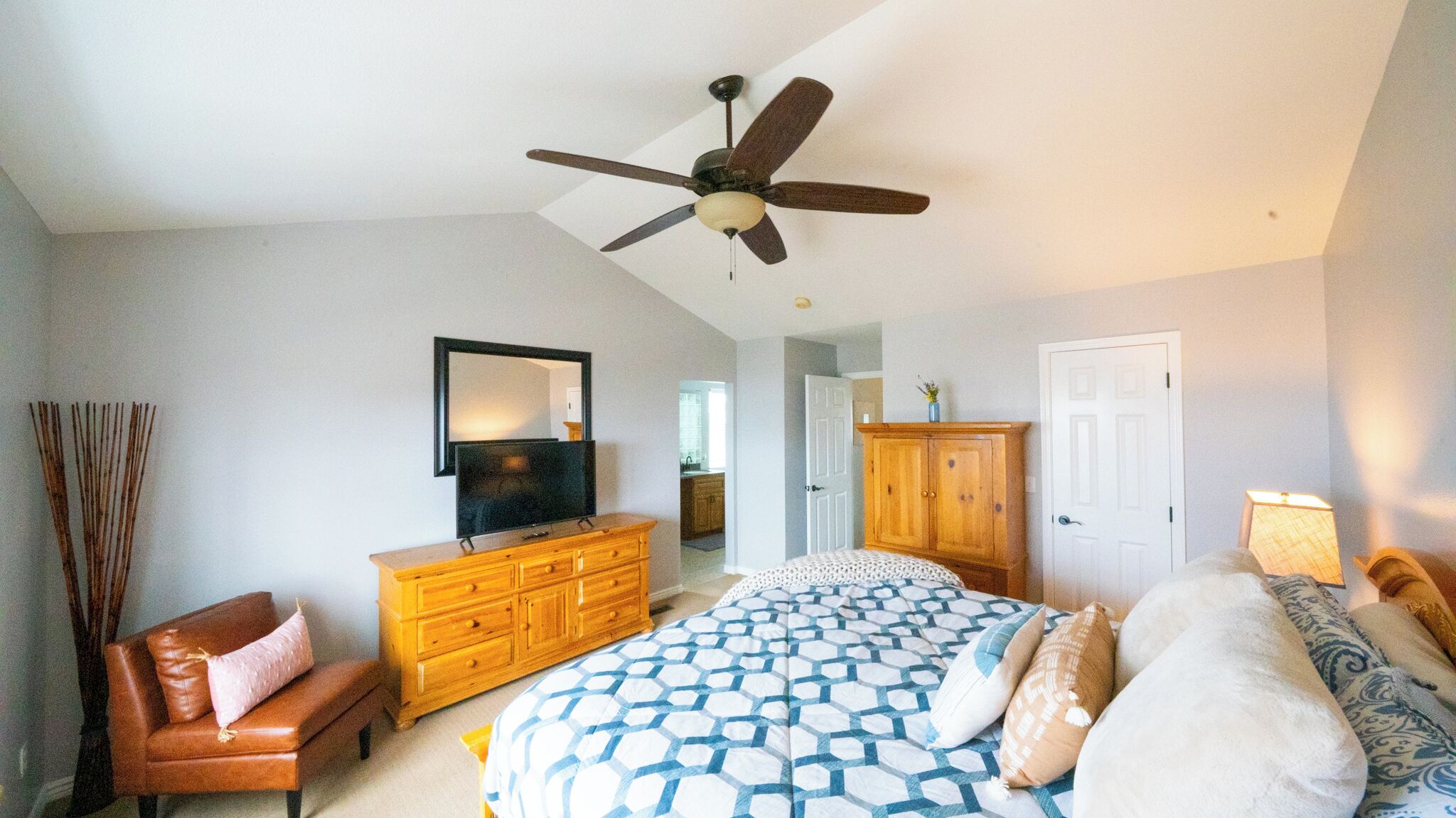 A bedroom with a bed, dresser, TV, ceiling fan, and a door leading to another room.