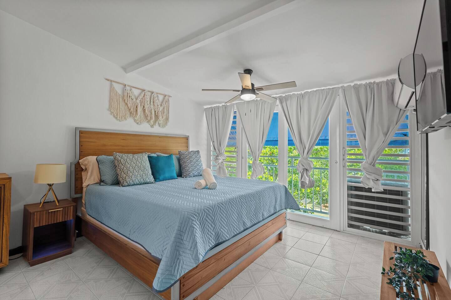 A bedroom with a bed, a nightstand, a ceiling fan, and a view of greenery through the window.