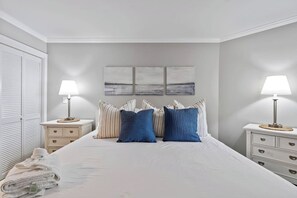 A bedroom with a bed, two bedside tables with lamps, a wall-mounted artwork, and a closet with a towel. - Ocean Creek Lodge 4 2276 - Beach, Pools, Hot Tubs (Myrtle Beach)