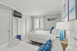 Ein Hotelzimmer mit zwei Betten, einem Fenster mit Jalousien, einem an der Wand befestigten Fernseher und einem kleinen Tisch mit Lampe.