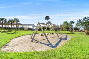 A playground with swings and a sandbox, surrounded by grass and trees. - Ocean Creek Lodge 4 2276 - Beach, Pools, Hot Tubs (Myrtle Beach)