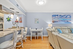 Eine moderne Küche mit weißen Schränken, ein Essbereich mit weißen Stühlen, ein beiges Sofa und ein Gemälde mit einer Strandansicht.