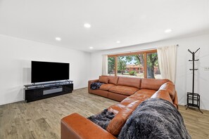 Un salon moderne avec un canapé d’angle, un téléviseur à écran plat et une grande fenêtre donnant sur l’extérieur.