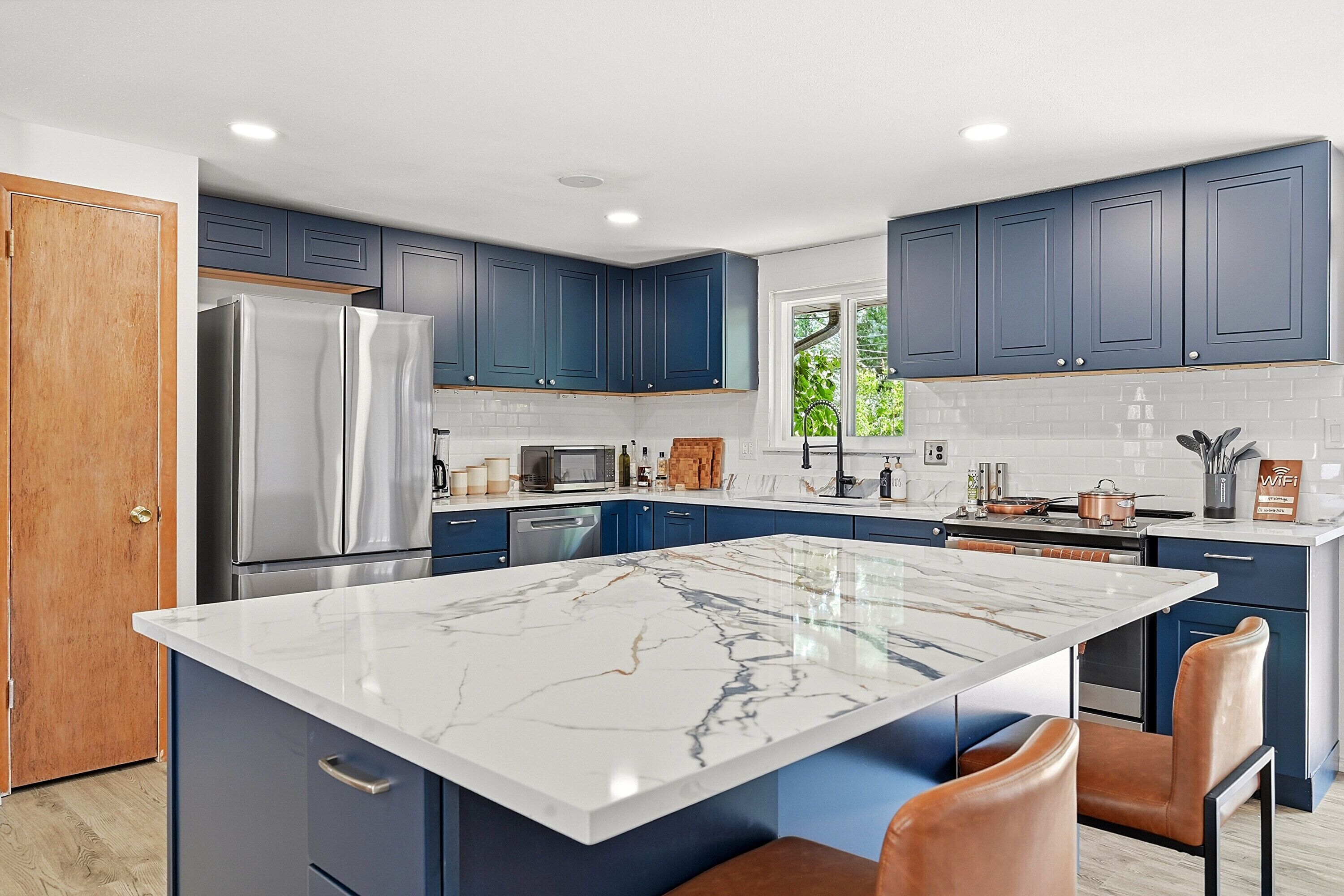 A modern kitchen with blue cabinets, a marble island, stainless steel appliances, and a view of greenery outside.