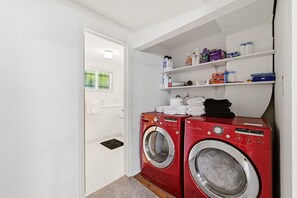 A laundry room with two red front-loading washing machines, a white door, and shelves above holding various cleaning supplies. - Peaceful 4-bedroom house with AC in Colorado Springs (Colorado Springs)
