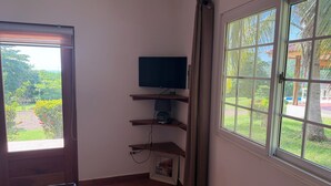 A room with a television on a wooden shelf, a window with a view of greenery, and a door leading outside. - Ocean view, with pool ,block to beach , , Cozy 1-room house in bucaro with AC (Tonosí)