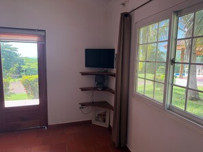 Una habitación con televisión sobre un estante de madera, una ventana con vistas a la vegetación y una puerta que da al exterior.