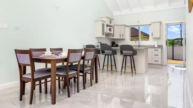 A modern kitchen with white cabinets, a dining area with wooden chairs, and a view of the outdoors through large windows. - DraxHall~3BR~Poo ~Beach Access - 24hr Security (St. Ann's Bay)