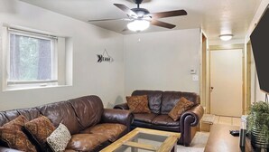 Un salon avec un canapé en cuir marron, une table basse en verre, un ventilateur de plafond et une porte visible au fond.