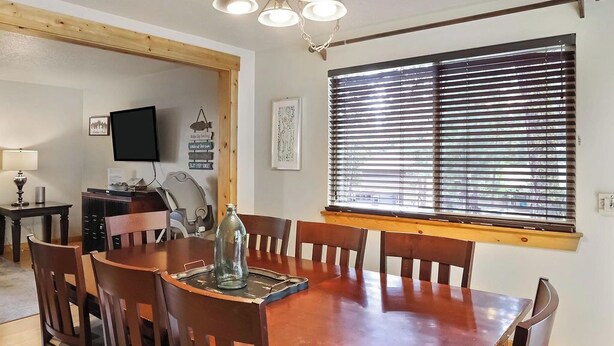 Un coin repas avec une table et des chaises en bois, un téléviseur et une fenêtre équipée de stores.