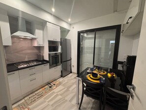 Cocina compacta con electrodomésticos modernos, comedor con mesa lista para dos personas y puerta corrediza de vidrio que da a un balcón.