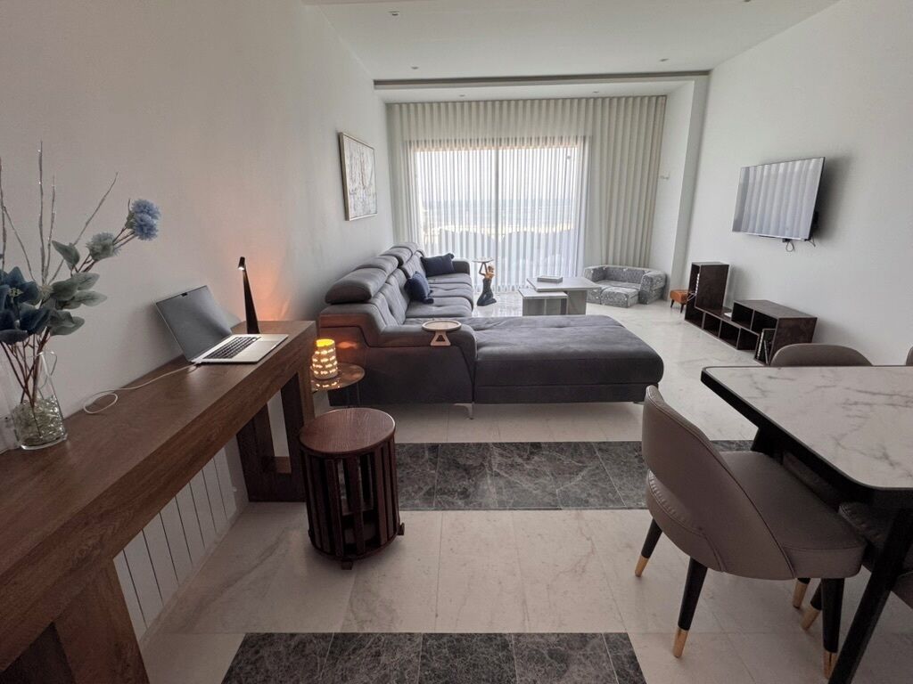 A modern living room with a sofa, a dining table, a TV mounted on the wall, and a laptop on a side table.