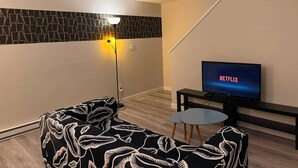 Un salon moderne avec un canapé, une petite table, un téléviseur affichant le logo de Netflix, un mur orné d’un motif géométrique et un escalier avec une touche de couleur bleue.