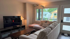 Un salon comprenant un canapé, une table basse, un téléviseur et une fenêtre donnant sur des maisons.