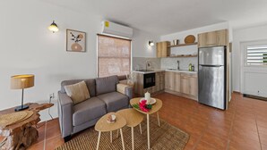 Uma sala moderna com um sofá cinza, uma mesa de centro e uma luminária. Uma cozinha adjacente possui uma geladeira de aço inoxidável, armários de madeira e uma janela com persianas.