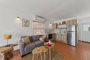 Ein modernes Wohnzimmer mit einem grauen Sofa, einem Couchtisch und einer Lampe. Daneben befindet sich eine Küche mit einem Edelstahlkühlschrank, Holzschränken und einem Fenster mit Jalousien.
