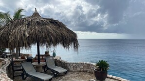 Área da piscina com espreguiçadeiras, um guarda-sol de palha e vista para o oceano.