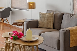 Ein Wohnzimmer mit einer Couch, einem kleinen runden Tisch mit Kerze und Blumen, einer Lampe und einem Sessel.