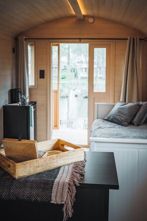 A cozy cabin interior with a bed, a small table with a tray, a black cabinet, and a window with curtains. - Camping Papillon Kinrooi - Pipowagen 4 p. terras by Interhome (Kinrooi)