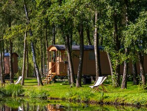 A row of wooden cabins with a small porch and a staircase, surrounded by trees and a calm body of water. - Camping Papillon Kinrooi - Pipowagen 4 p. terras by Interhome (Kinrooi)
