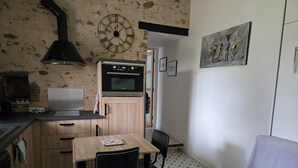 Eine kompakte Küche mit Mikrowelle, Backofen und Holzschränken. Ein Esstisch mit Stühlen und eine Couch mit einem Blumenkissen bieten Komfort. Eine Uhr und Kunstwerke zieren die Wände.