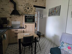 A compact kitchen with a microwave, oven, and wooden cabinets. There's a dining table with chairs and a couch with a floral cushion. A clock and artwork adorn the walls. - A charming, quiet, and rustic little cottage with a small garden and private parking. (Clisson)