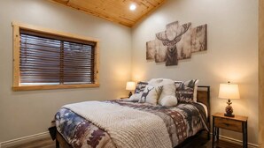 A bedroom with a bed, a window with blinds, a wall with a deer painting, and a nightstand with a lamp. - Hilltop Cabin | Fireplace - Games - Views (Big Bear)