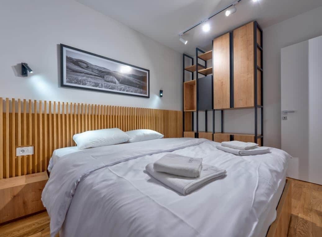 A modern hotel room with a large bed, wooden headboard, a mounted picture, built-in shelving, and a bedside table.
