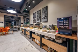 Montaje de bufé con diversos alimentos, una máquina de café y mesas con sillas.