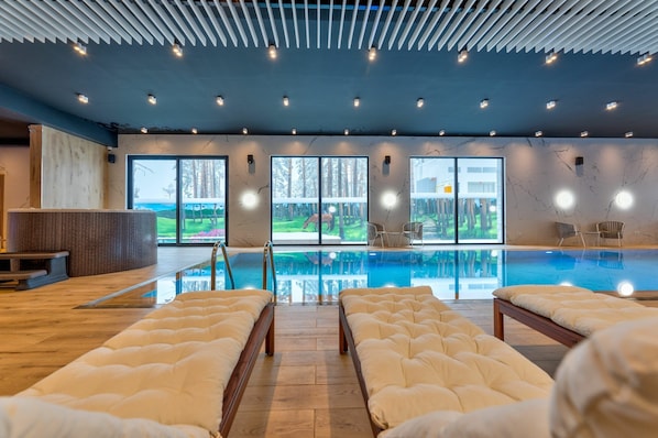 A modern indoor swimming pool with clear glass walls, wooden flooring, and multiple seating areas with cushioned loungers. - Konak Apartmani & Spa (Zlatibor)