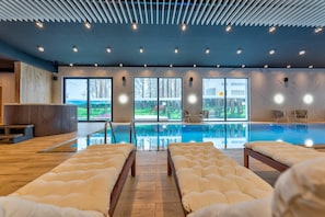 Una piscina interior moderna con paredes de vidrio transparente, suelo de madera y varias zonas de estar con sillones reclinables acolchados.