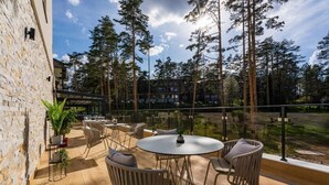 Een terras buiten met ronde tafels en rieten stoelen, omgeven door hoge bomen en een stenen muur.
