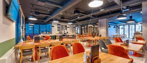 Un espace de bureau moderne doté de plusieurs tables en bois et de chaises orange, de grandes fenêtres et d’un éclairage de style industriel.