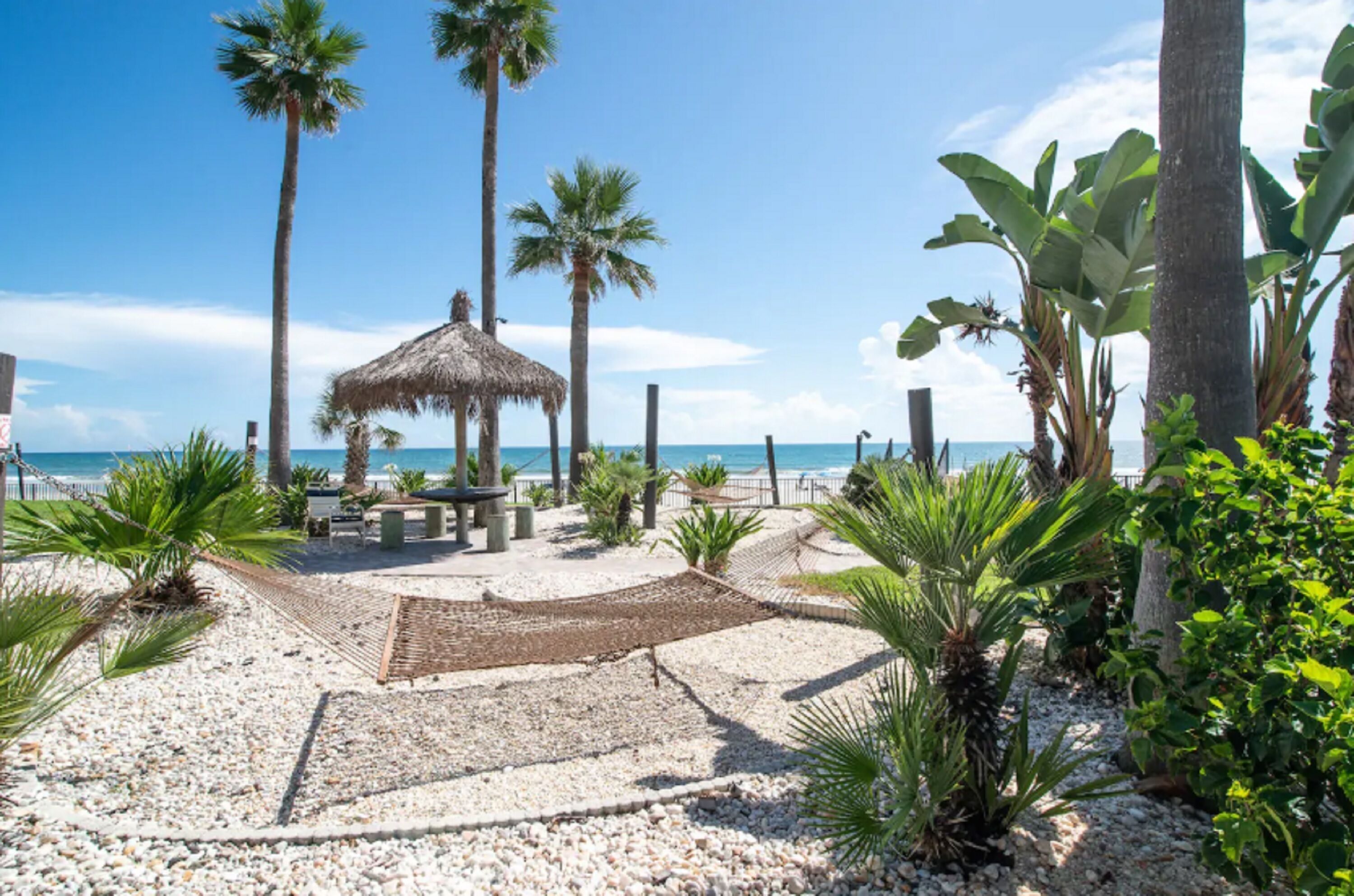 Ein Strandbereich mit einem strohgedeckten Pavillon, Palmen und einem Kiesweg, der zum Meer führt.