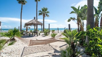 Ein Strandbereich mit einem strohgedeckten Pavillon, Palmen und einem Kiesweg, der zum Meer führt.