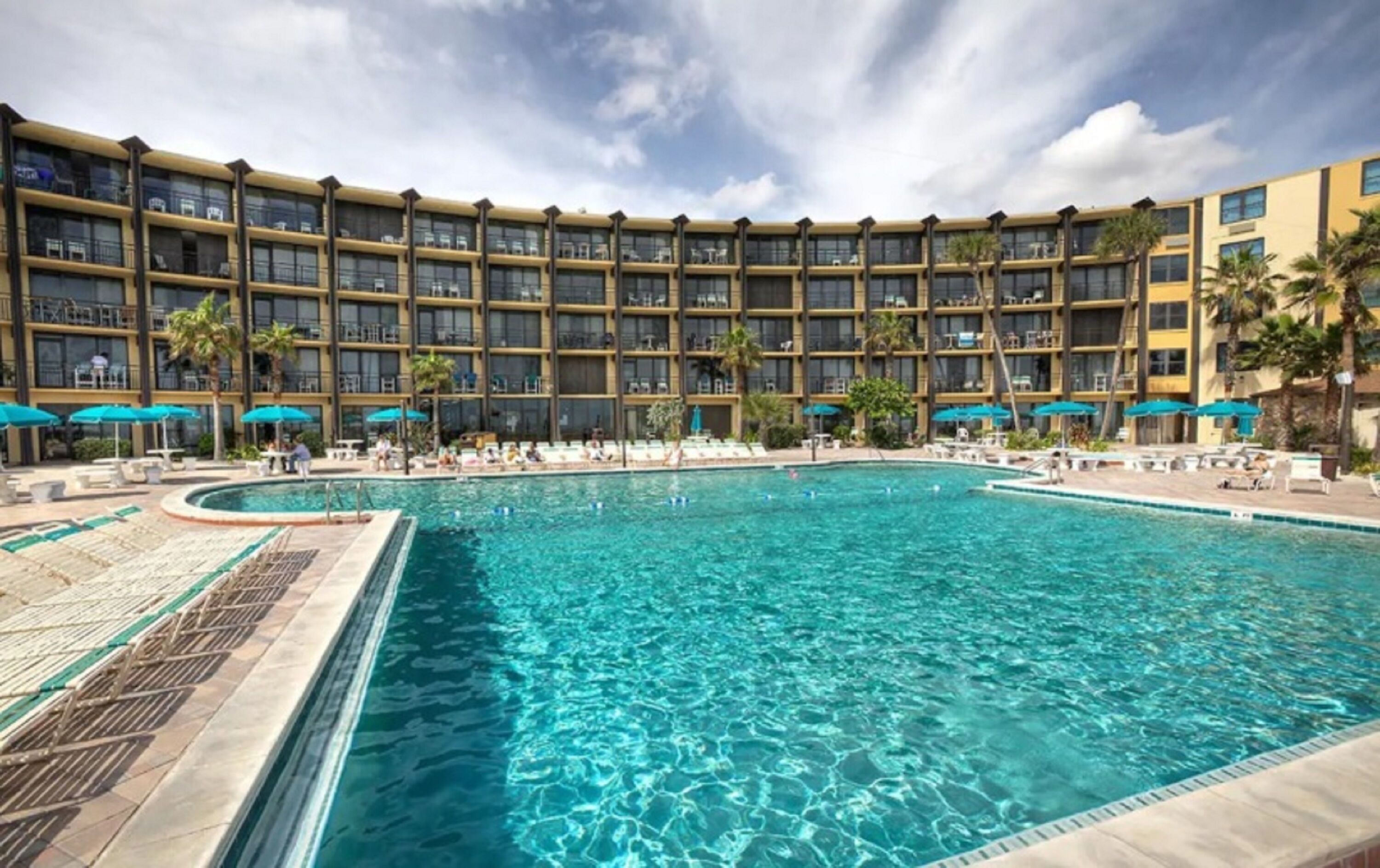 Ein Hotel mit einem großen Pool, umgeben von Liegestühlen und Sonnenschirmen, mehreren Etagen mit Balkonen und einem wolkenlosen Himmel.